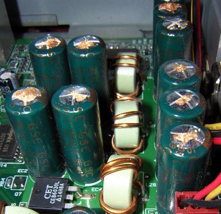 Figure 8 — Bulged and vented aluminium electrolytics on a PC board, the visual signature of the "capacitor plague": domed tops and split vents. Source: Jens Both, Wikimedia Commons, CC BY-SA 3.0.