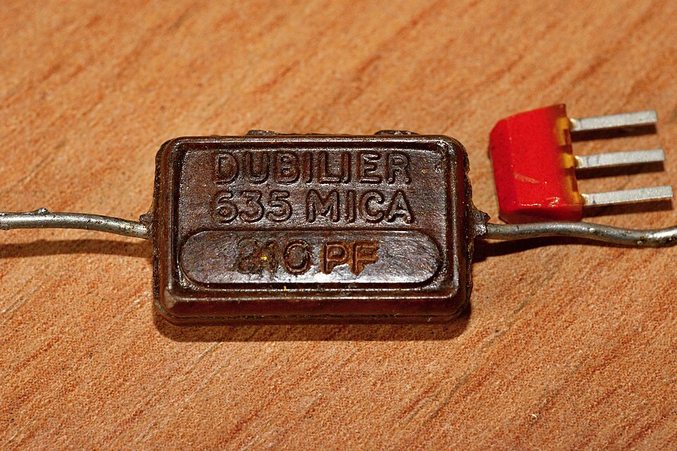 Figure 7 — A Dubilier "postage-stamp" moulded-mica capacitor of the 1940s. Mica's stable, low-loss dielectric made it the precision choice for RF and timing before ceramics matured. The coloured-do…